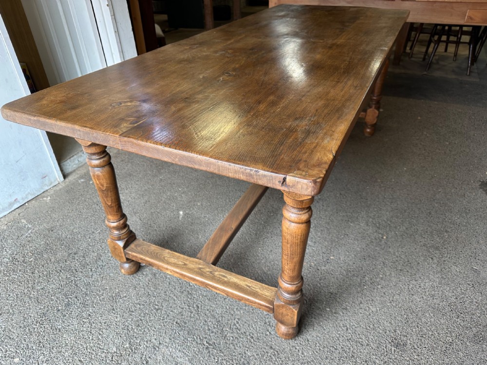 A long rectangular antique elm dining table featuring turned wooden legs connected by a central stretcher base and a smooth timber top. View VI