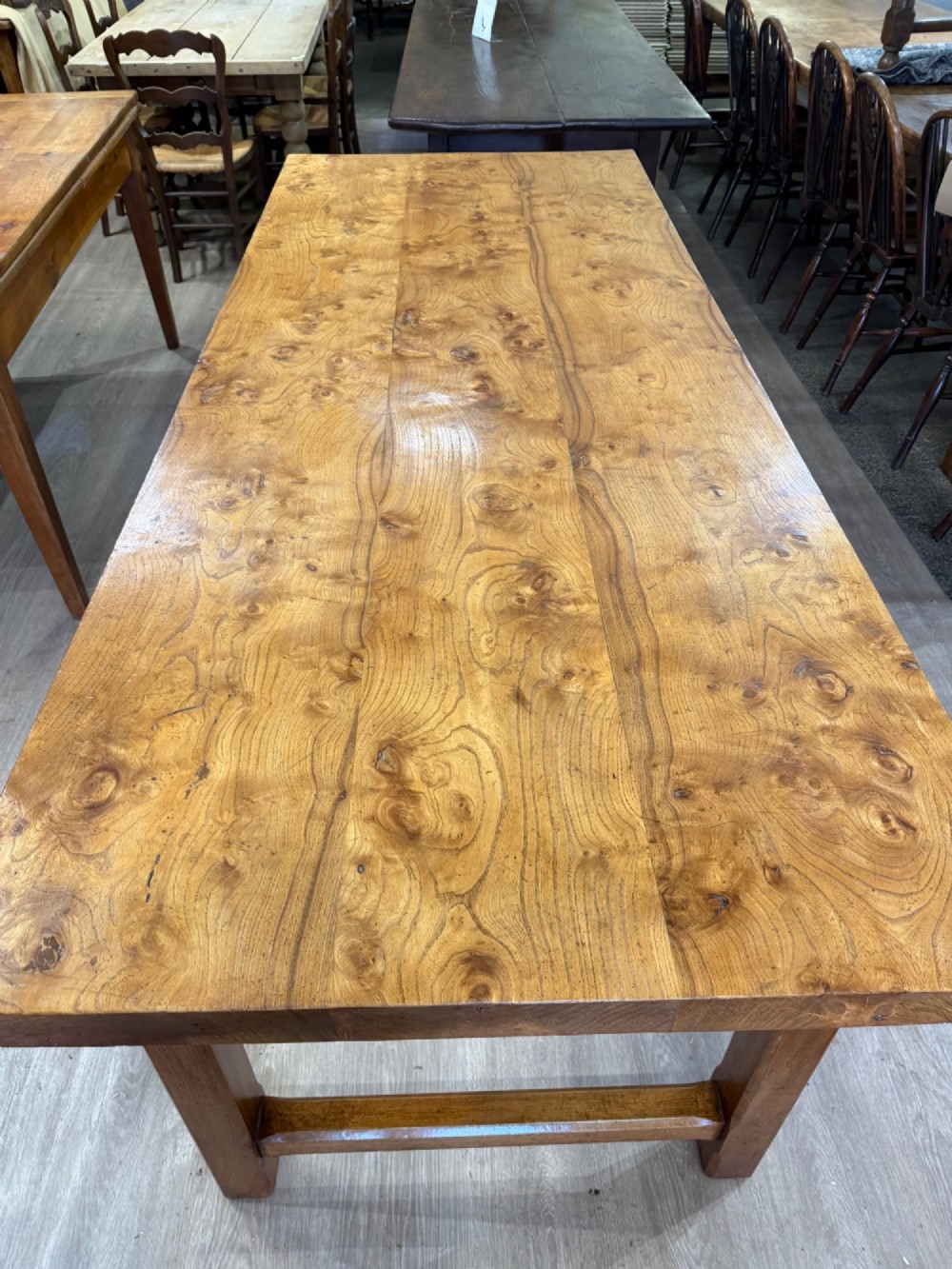 A long rectangular wooden farmhouse dining table featuring a plank top and sturdy trestle leg base in a warm honey oak finish. View IV
