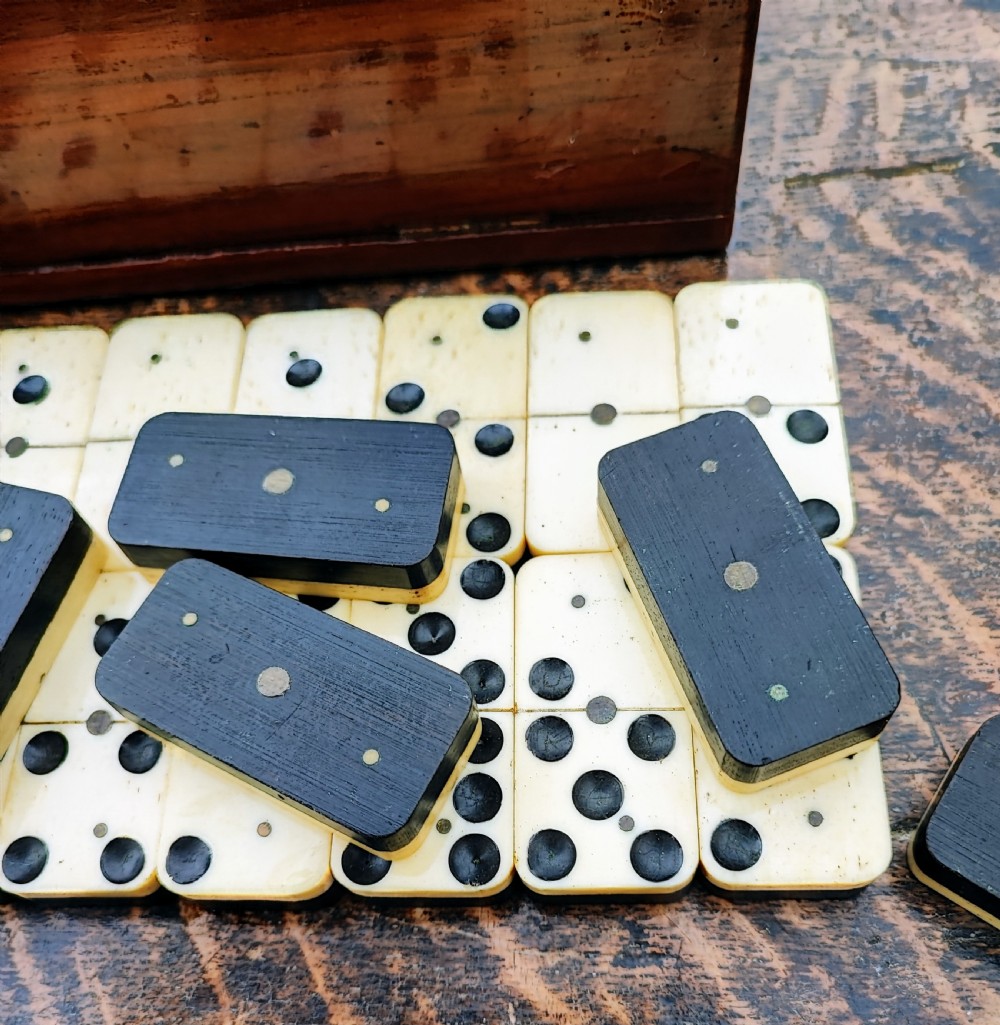 A full set of Victorian bone and ebony dominoes arranged in rows on a wooden surface in front of a fitted rectangular wooden box. View II
