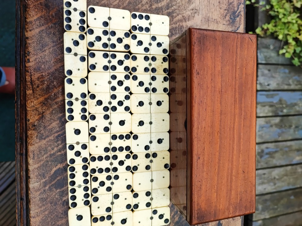 A full set of Victorian bone and ebony dominoes arranged in rows on a wooden surface in front of a fitted rectangular wooden box. View III