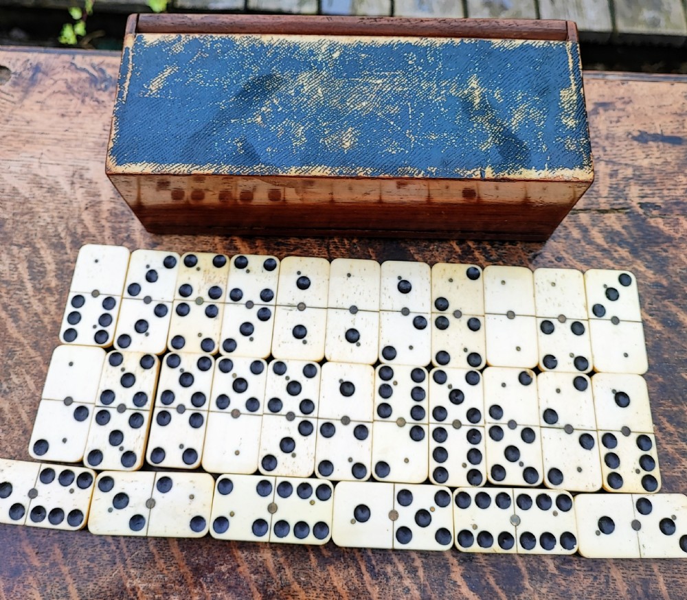 A full set of Victorian bone and ebony dominoes arranged in rows on a wooden surface in front of a fitted rectangular wooden box. View VI