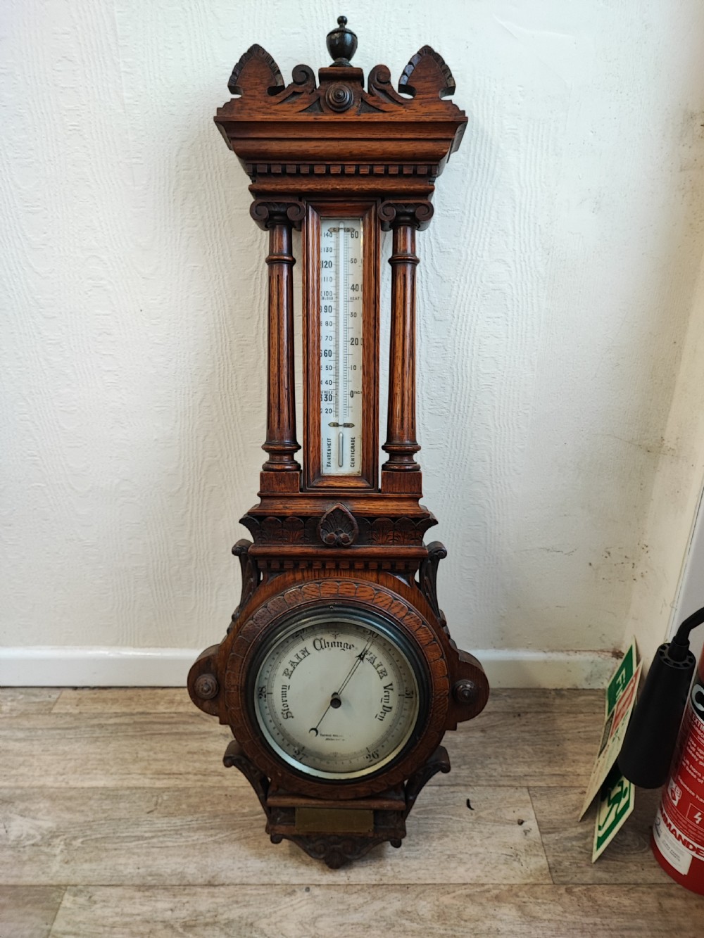 A carved oak wall barometer featuring an upper thermometer flanked by turned columns above a circular aneroid dial.