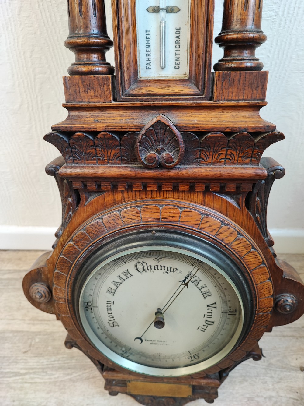 A carved oak wall barometer featuring an upper thermometer flanked by turned columns above a circular aneroid dial. View VII