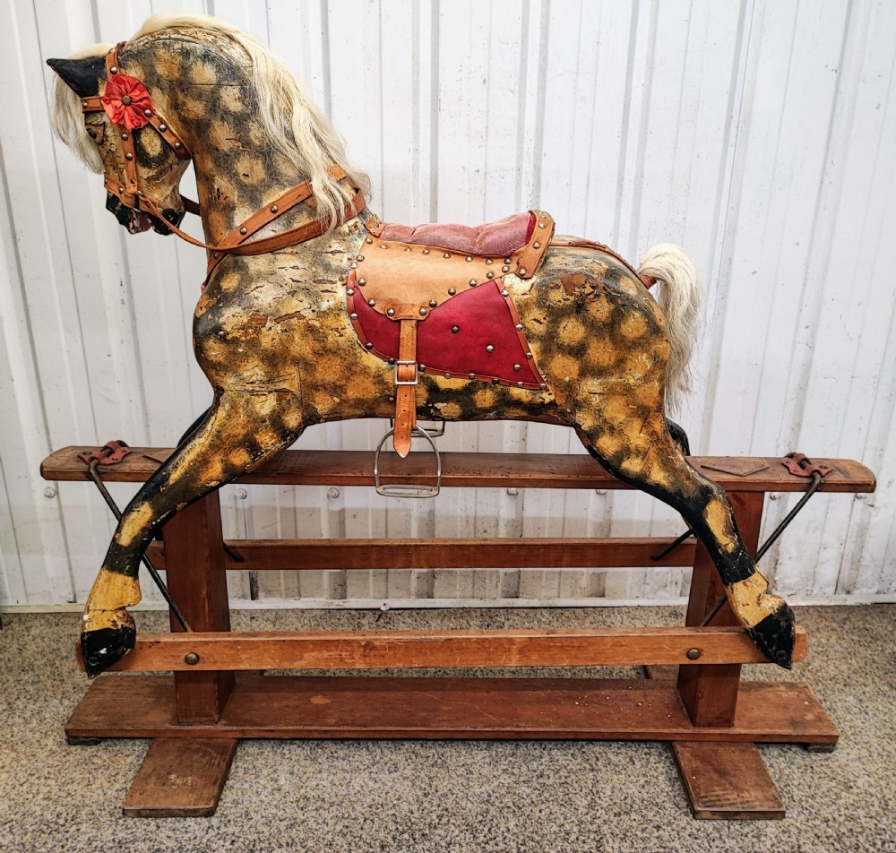 A large antique carved wooden rocking horse with a dappled painted finish, leather saddle, horsehair mane, and tail, mounted on a wooden safety stand.
