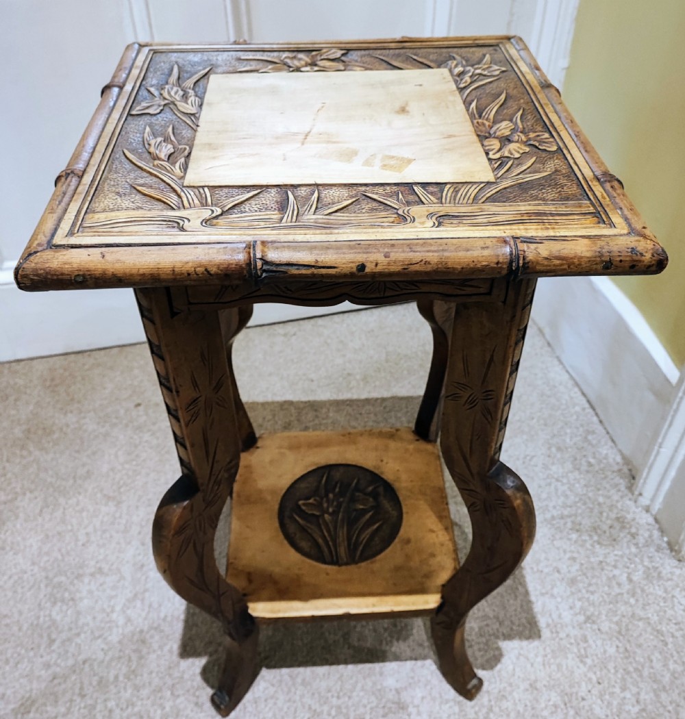 A square Arts and Crafts wooden side table featuring carved iris botanical motifs, bamboo-style edge moulding, shaped legs, and a carved lower shelf.