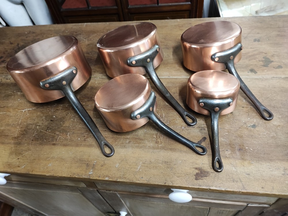 A set of five graduated antique copper saucepans with dark cast iron handles arranged upside down.