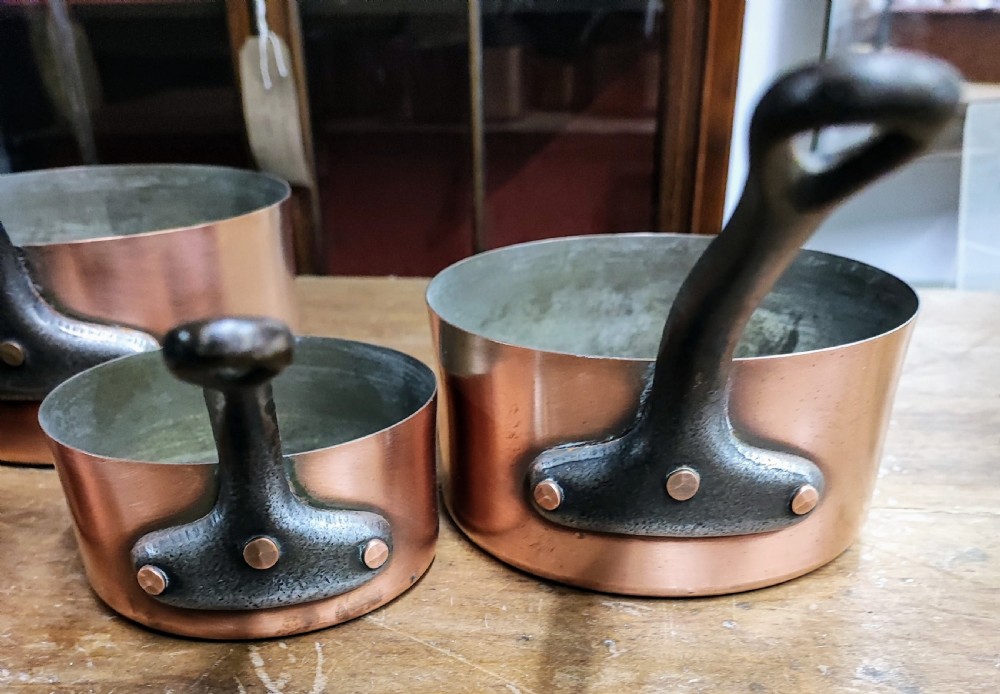 A set of five graduated antique copper saucepans with dark cast iron handles arranged upside down. View III