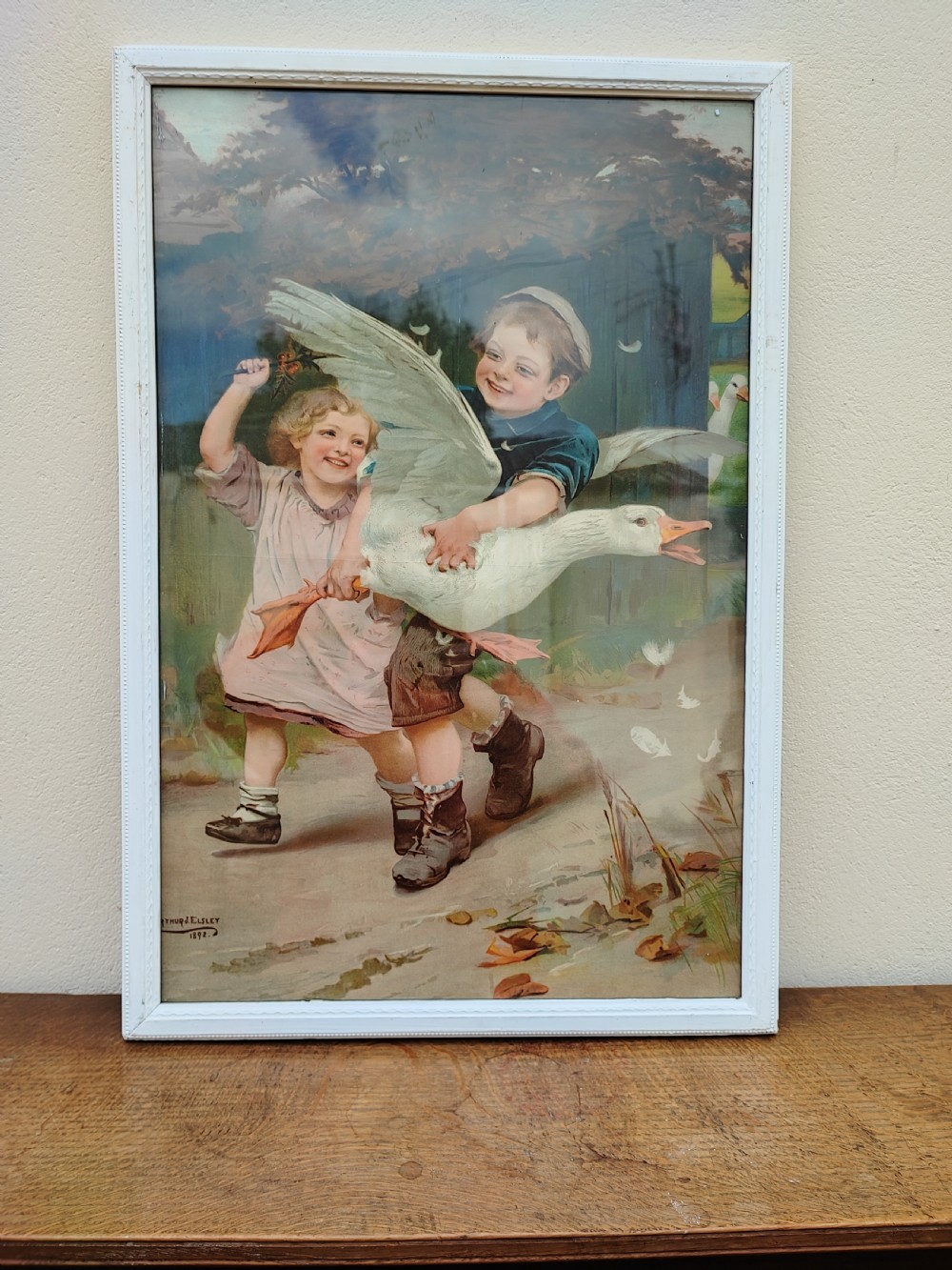 A framed Victorian chromolithograph print depicting a young boy holding a goose alongside a smiling young girl in a narrow white frame.