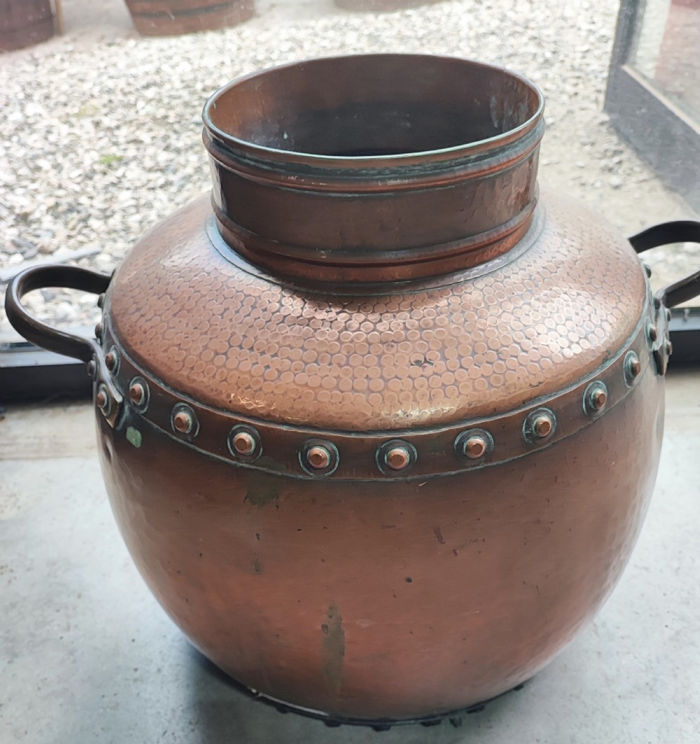A large, rounded Arts and Crafts copper vessel featuring a hand-hammered shoulder, riveted metal band, and dual side handles.
