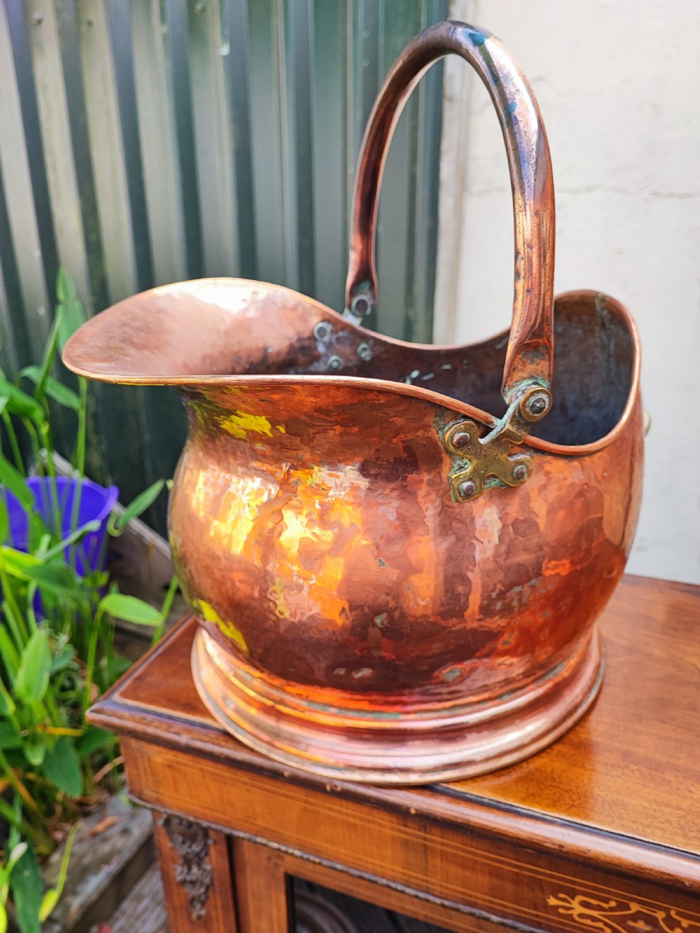 A helmet-shaped antique copper coal scuttle featuring an arched metal handle secured with brass rivets over a round stepped base.