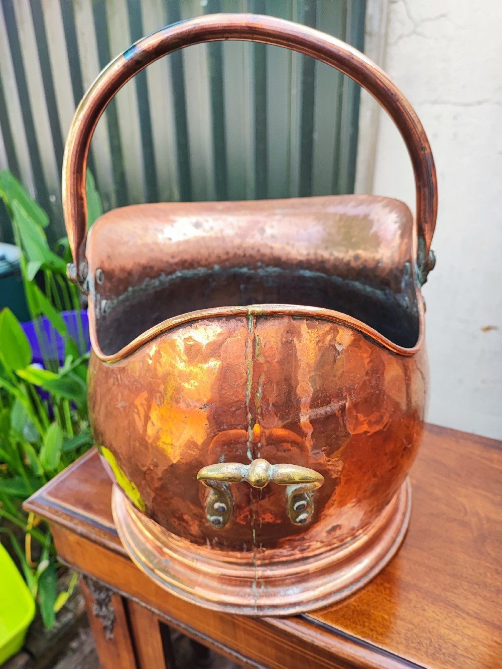 A helmet-shaped antique copper coal scuttle featuring an arched metal handle secured with brass rivets over a round stepped base. View III