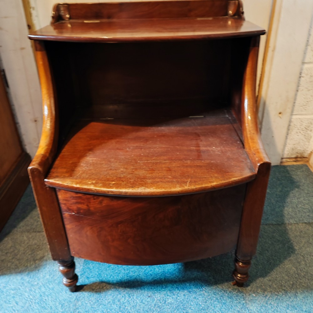 An antique Regency flame mahogany bedside commode with a top shelf tier, a stepped middle surface, and a curved front raised on turned feet.