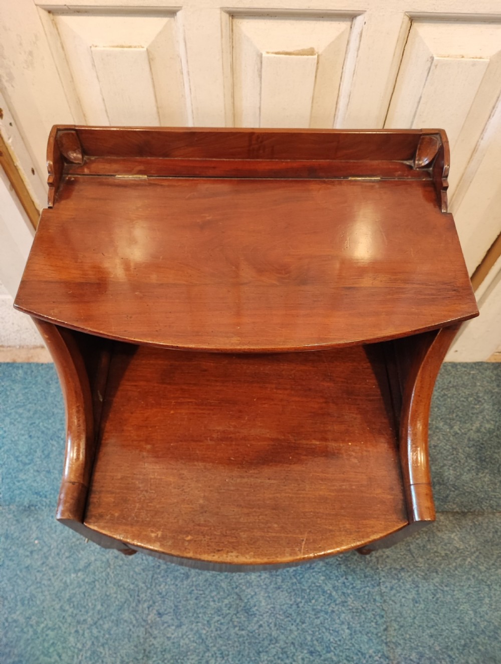 An antique Regency flame mahogany bedside commode with a top shelf tier, a stepped middle surface, and a curved front raised on turned feet. View III