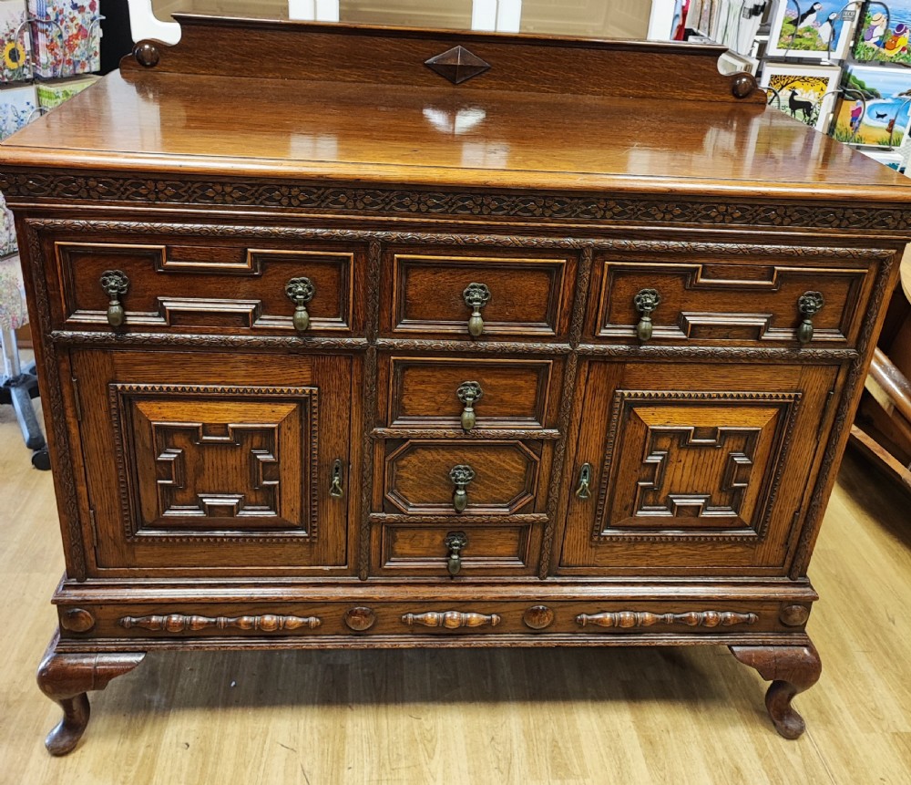 A carved dark oak sideboard featuring geometric panelled cupboard doors, multiple drawers with brass drop handles, cabriole legs, and a low upstand.