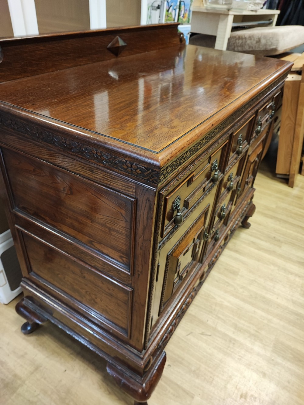 A carved dark oak sideboard featuring geometric panelled cupboard doors, multiple drawers with brass drop handles, cabriole legs, and a low upstand. View VI