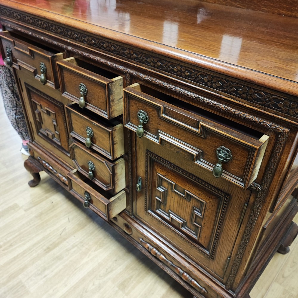 A carved dark oak sideboard featuring geometric panelled cupboard doors, multiple drawers with brass drop handles, cabriole legs, and a low upstand. View XIV