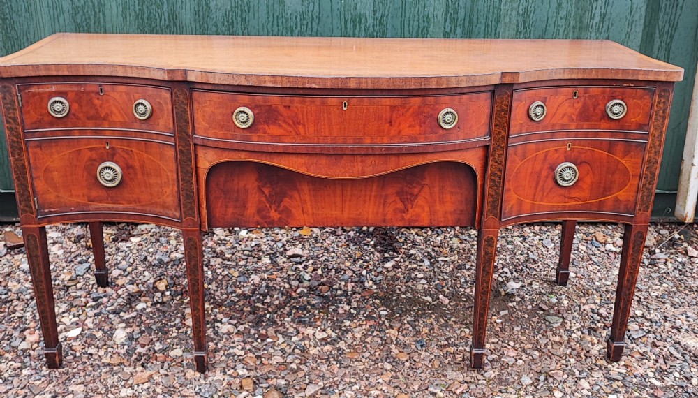 A George III style serpentine mahogany sideboard featuring marquetry inlay, brass ring handles, an arched central drawer, and square tapered legs.
