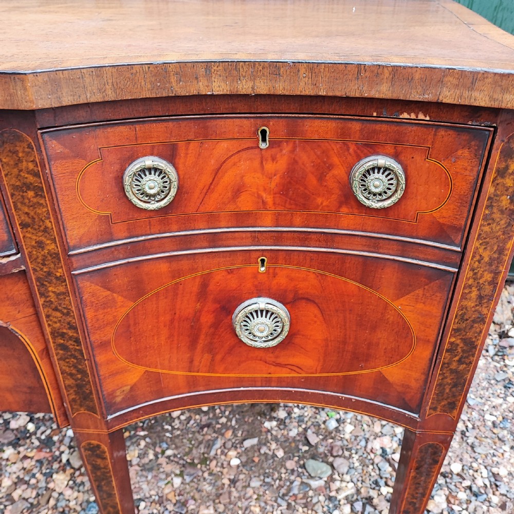 A George III style serpentine mahogany sideboard featuring marquetry inlay, brass ring handles, an arched central drawer, and square tapered legs. View II