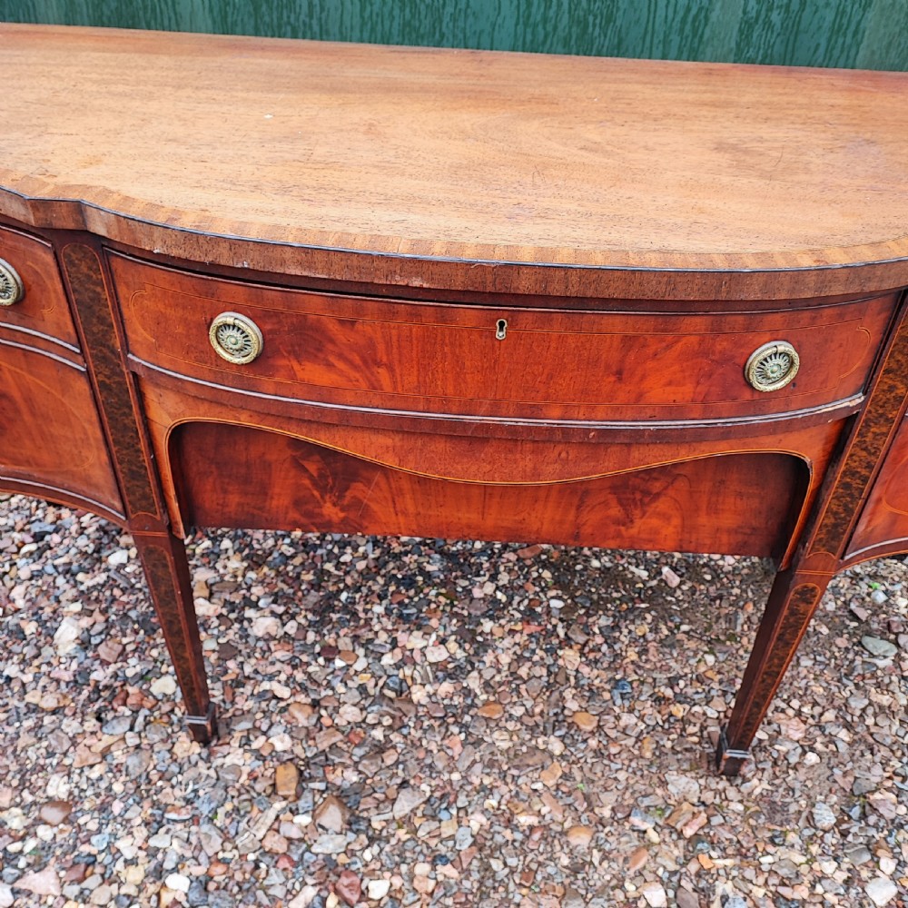 A George III style serpentine mahogany sideboard featuring marquetry inlay, brass ring handles, an arched central drawer, and square tapered legs. View X