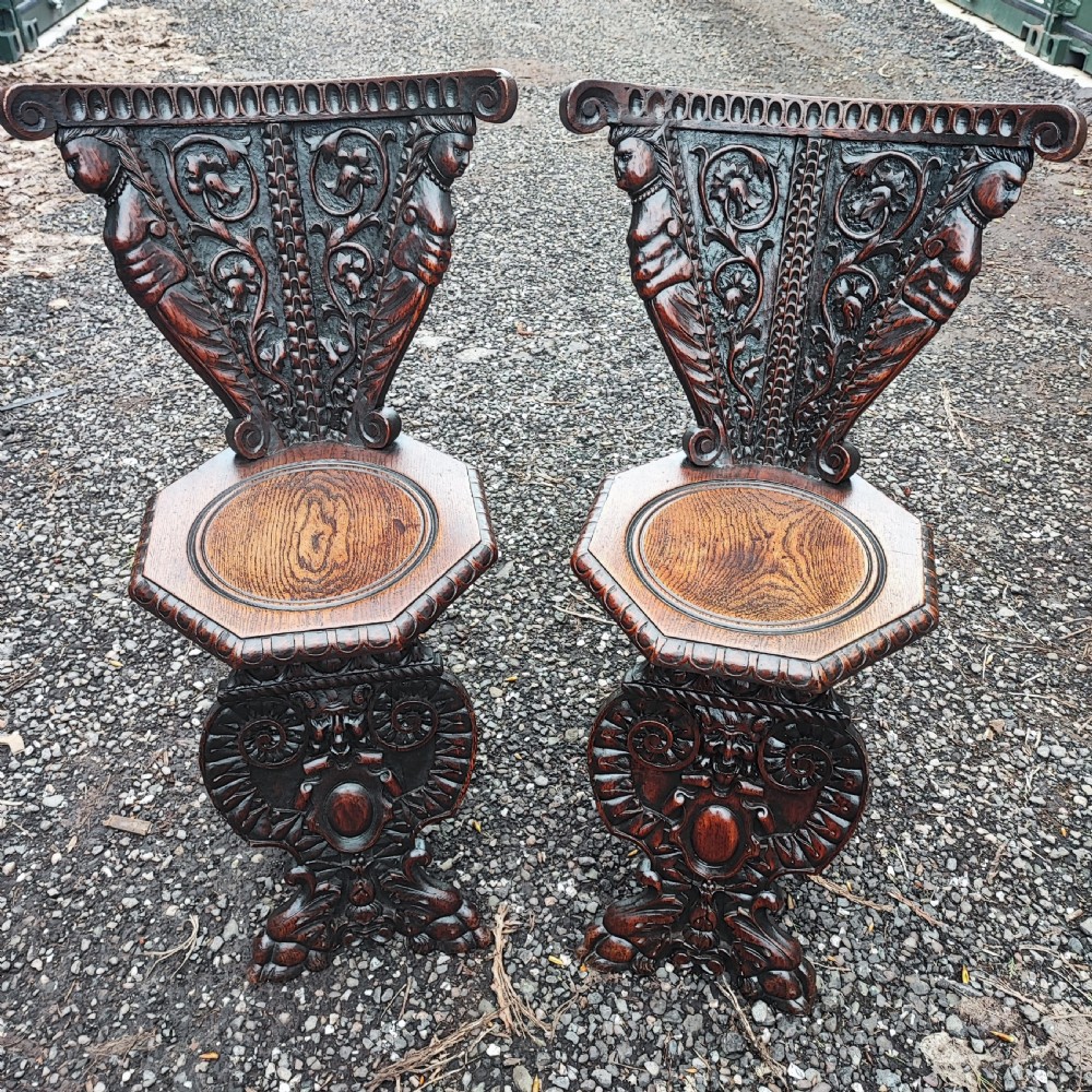 A pair of dark oak sgabello hall chairs featuring heavily carved fan-shaped backrests with figural details, octagonal seats, and ornate trestle bases.