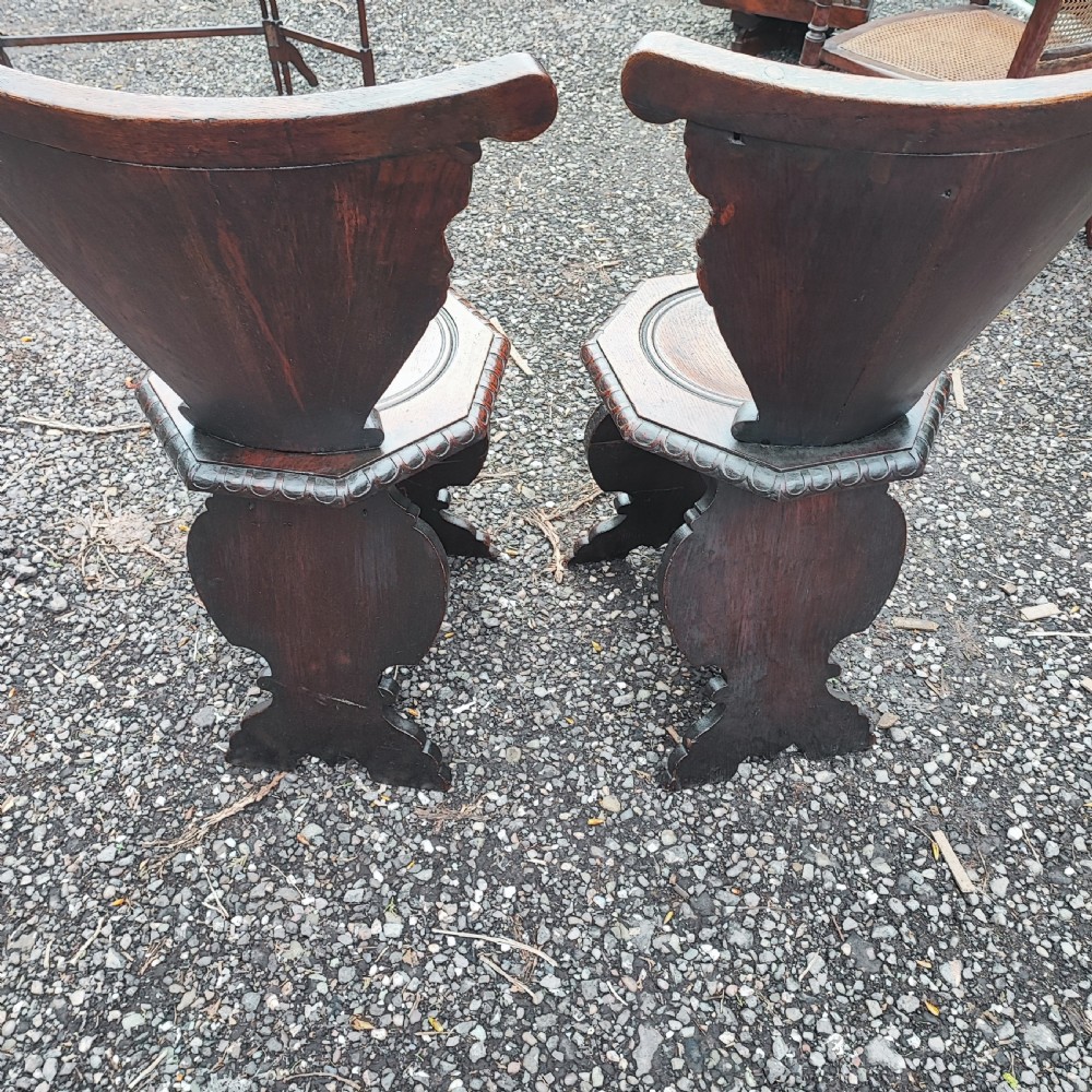 A pair of dark oak sgabello hall chairs featuring heavily carved fan-shaped backrests with figural details, octagonal seats, and ornate trestle bases. View II