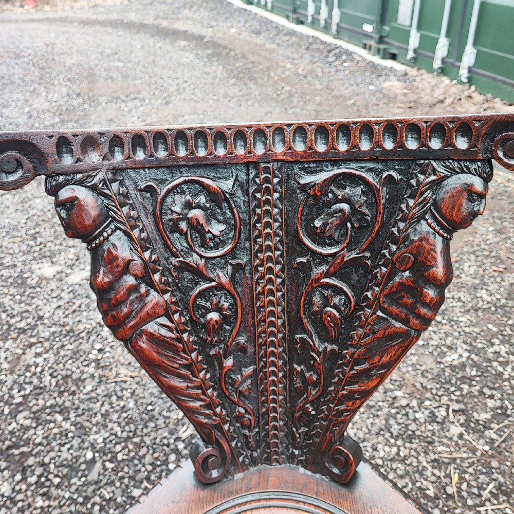 A pair of dark oak sgabello hall chairs featuring heavily carved fan-shaped backrests with figural details, octagonal seats, and ornate trestle bases. View III