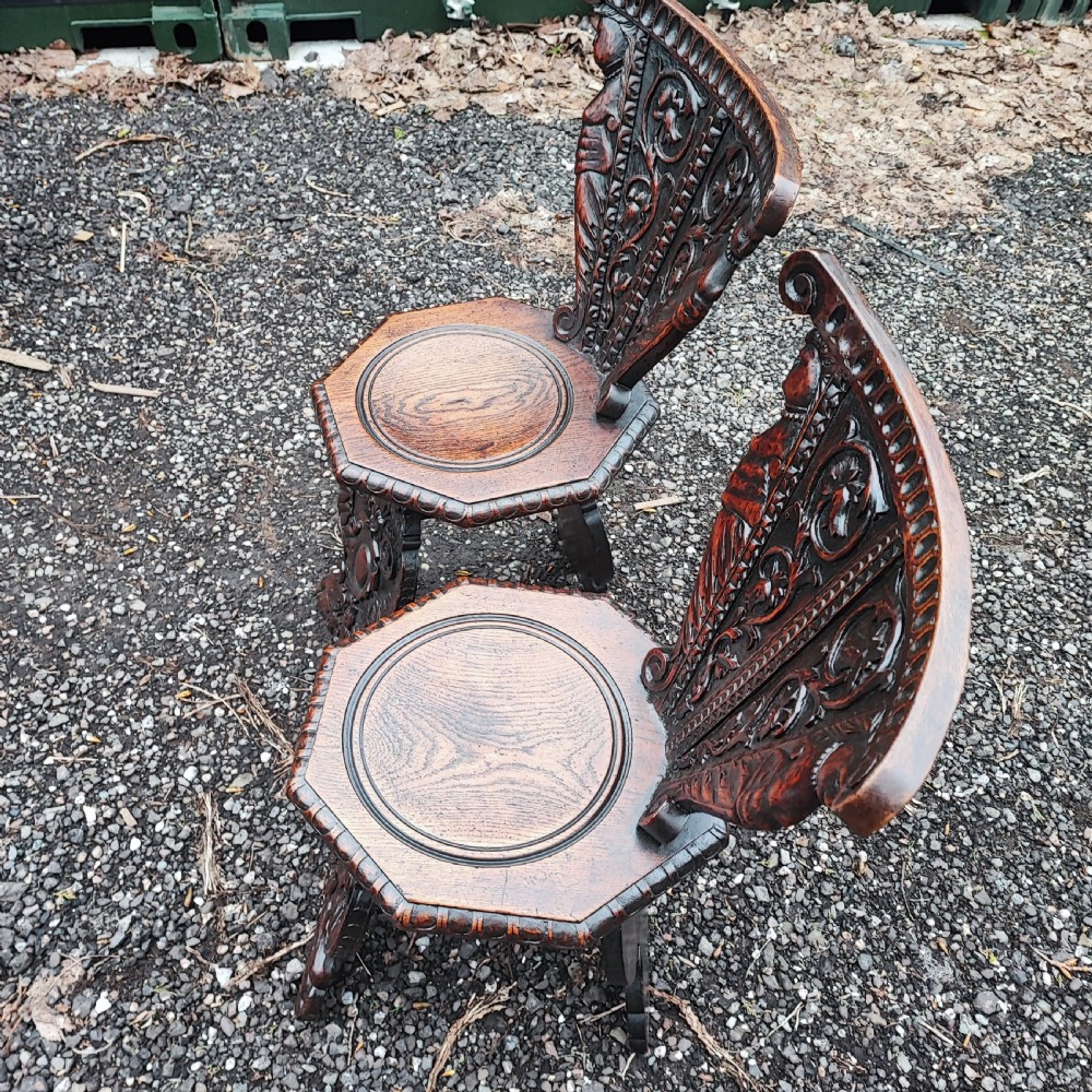 A pair of dark oak sgabello hall chairs featuring heavily carved fan-shaped backrests with figural details, octagonal seats, and ornate trestle bases. View V