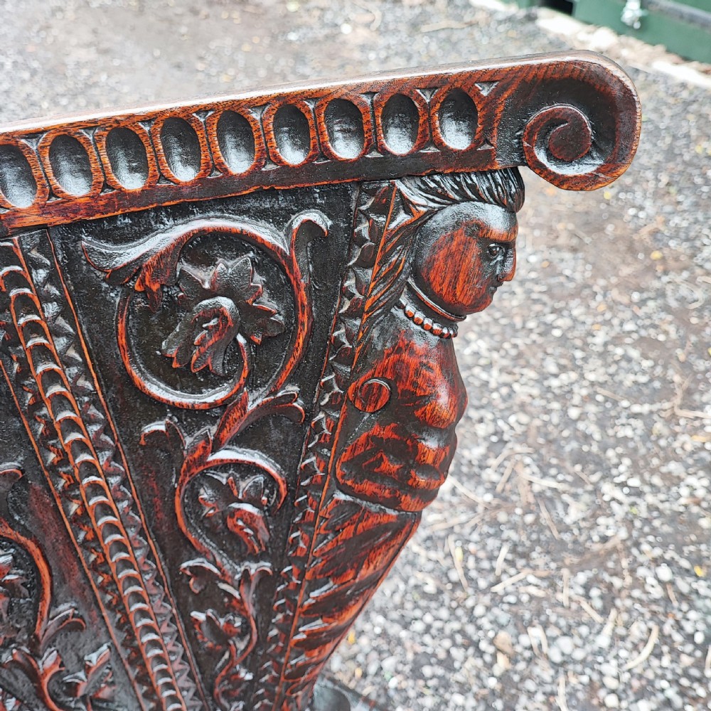 A pair of dark oak sgabello hall chairs featuring heavily carved fan-shaped backrests with figural details, octagonal seats, and ornate trestle bases. View VII