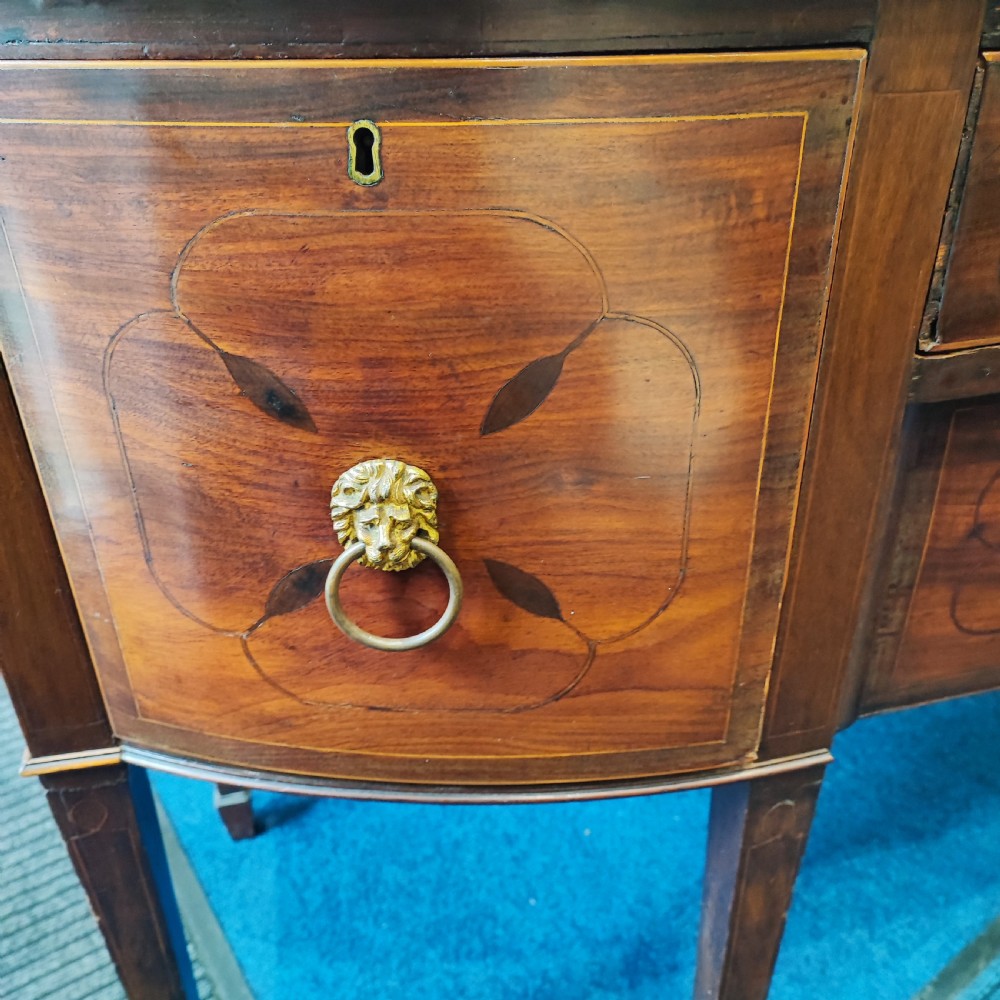 A Regency mahogany bowfront sideboard featuring inlaid floral motifs, brass lion-mask ring handles, and tapered square legs. View III