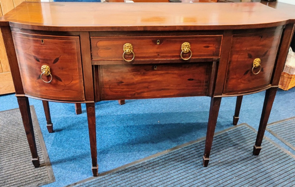A Regency mahogany bowfront sideboard featuring inlaid floral motifs, brass lion-mask ring handles, and tapered square legs. View IV