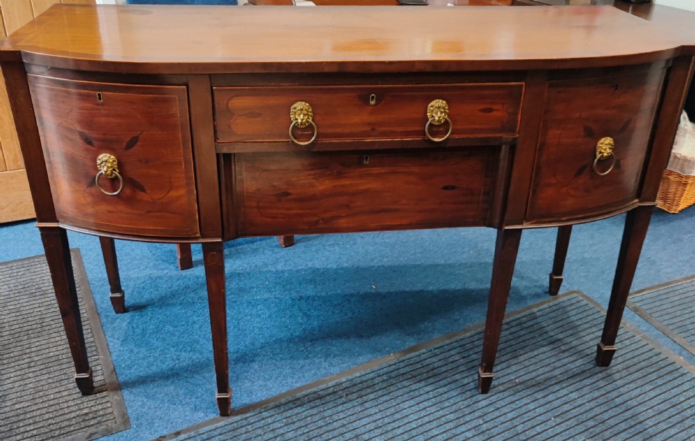 A Regency mahogany bowfront sideboard featuring inlaid floral motifs, brass lion-mask ring handles, and tapered square legs. View V