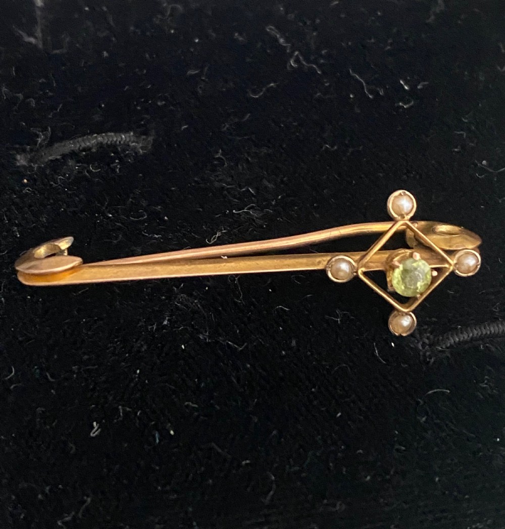 An antique yellow gold safety pin brooch featuring a central geometric diamond motif set with a round green peridot and four small split pearls.