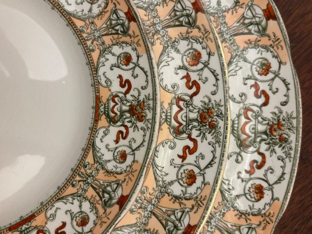 An extensive antique Doulton dinner service featuring multiple ceramic plates, side dishes, and covered tureens decorated with a scrolled terracotta and gilt border pattern. View II