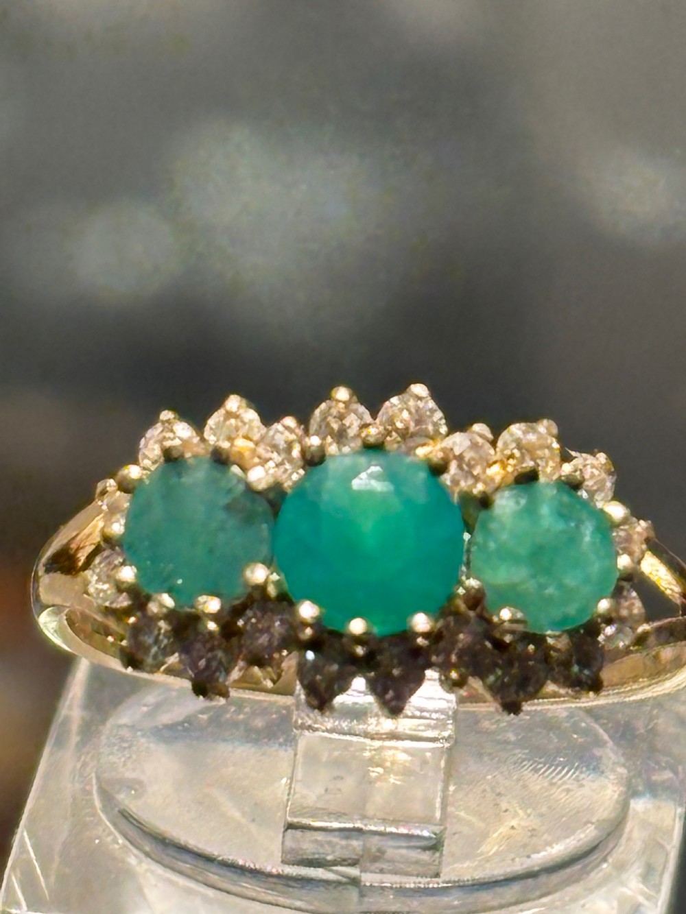 A vintage gold ring featuring three central emeralds surrounded by a halo of small diamonds, resting inside a black velvet jewellery box. View IX