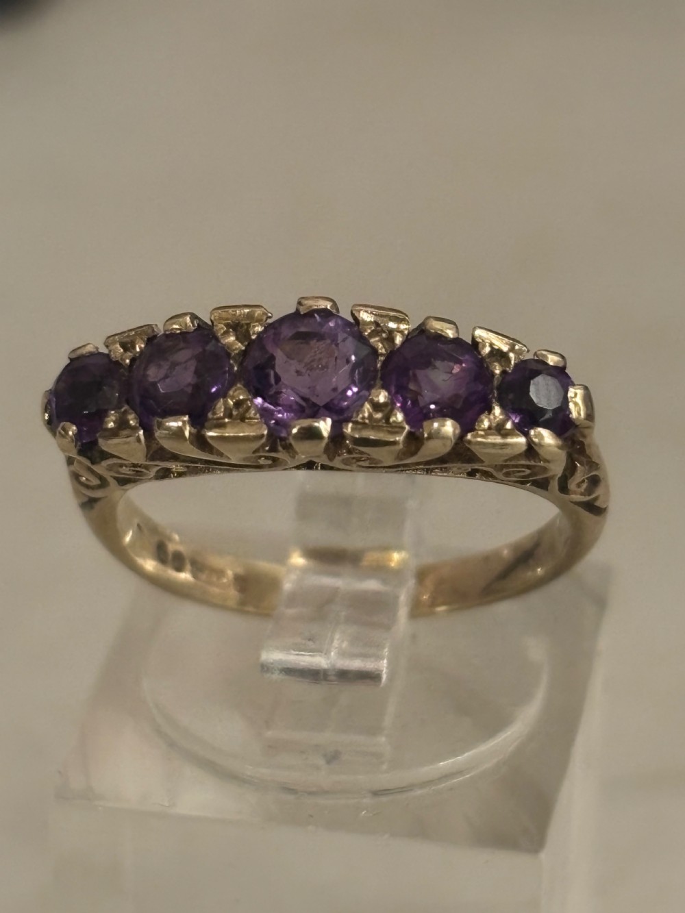 A yellow gold five stone ring set with five graduated round purple amethyst gemstones and carved scrollwork on the side gallery.