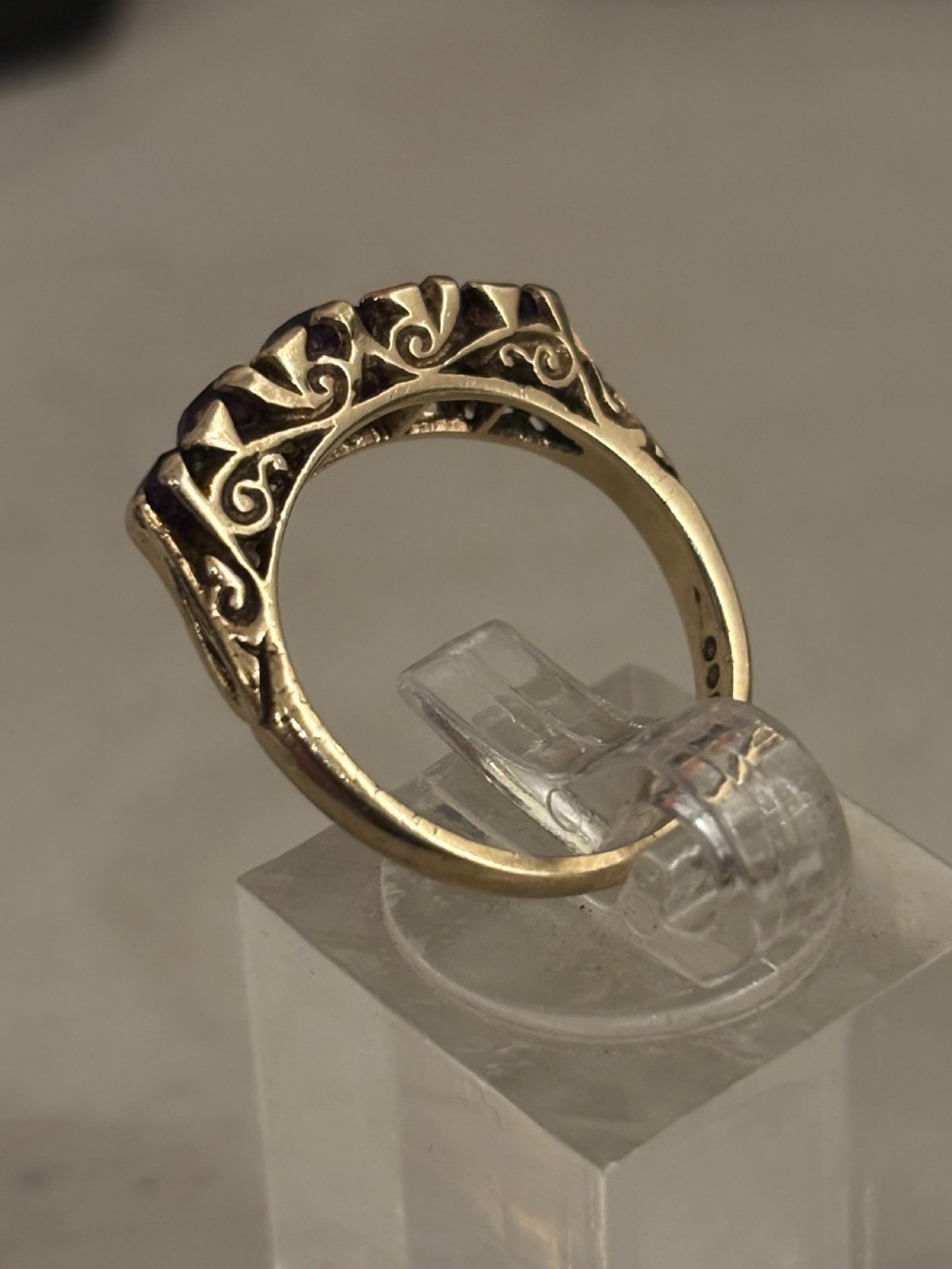 A yellow gold five stone ring set with five graduated round purple amethyst gemstones and carved scrollwork on the side gallery. View II