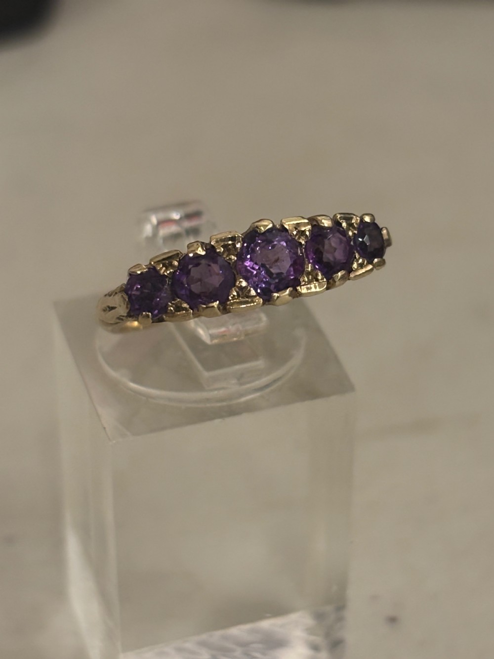 A yellow gold five stone ring set with five graduated round purple amethyst gemstones and carved scrollwork on the side gallery. View V