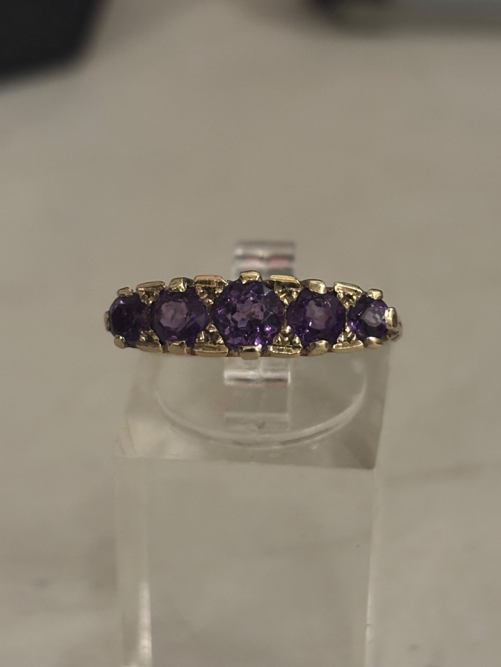 A yellow gold five stone ring set with five graduated round purple amethyst gemstones and carved scrollwork on the side gallery. View VI