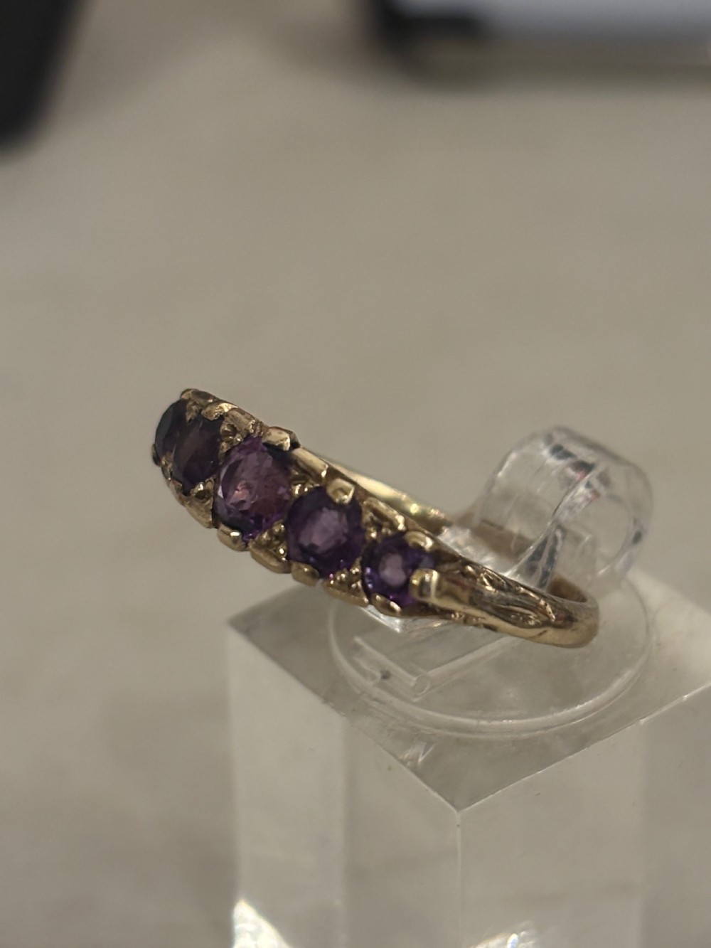 A yellow gold five stone ring set with five graduated round purple amethyst gemstones and carved scrollwork on the side gallery. View VII