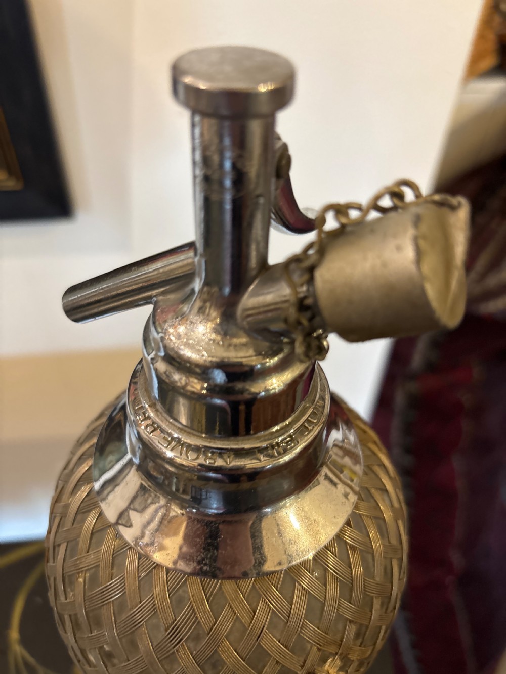 A 1930s glass soda syphon featuring a protective woven metal mesh casing and a polished chrome dispensing head with a chained stopper. View III