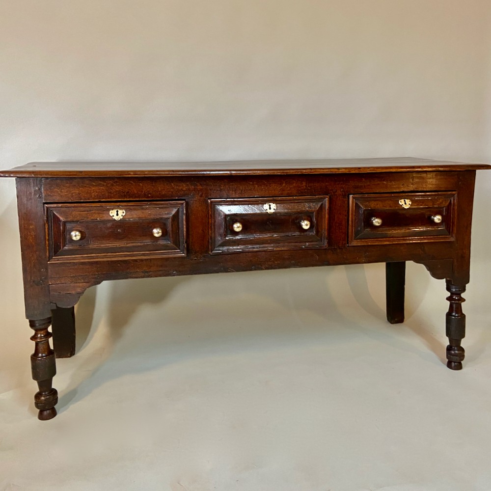 A late 17th-century dark oak low dresser featuring three geometric panelled drawers with brass escutcheons and turned front legs.