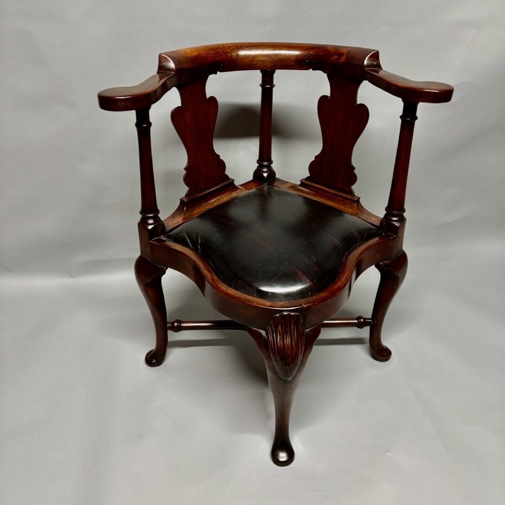 An 18th-century George III dark mahogany corner chair featuring a curved top rail, twin shaped splats, a dark leather seat, turned stretchers, and a front cabriole leg carved with a shell motif.