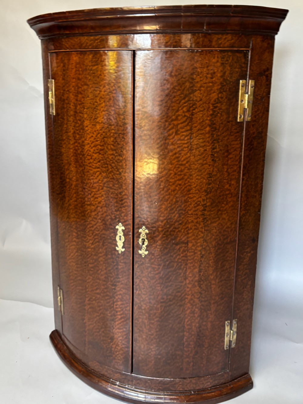 A Georgian bowfront mahogany corner cupboard featuring distinctive plum pudding grain, brass escutcheons, and visible brass H-hinges.
