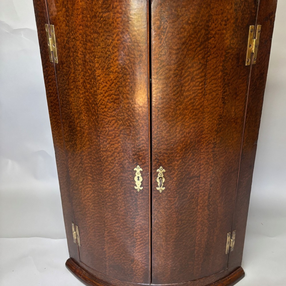 A Georgian bowfront mahogany corner cupboard featuring distinctive plum pudding grain, brass escutcheons, and visible brass H-hinges. View II
