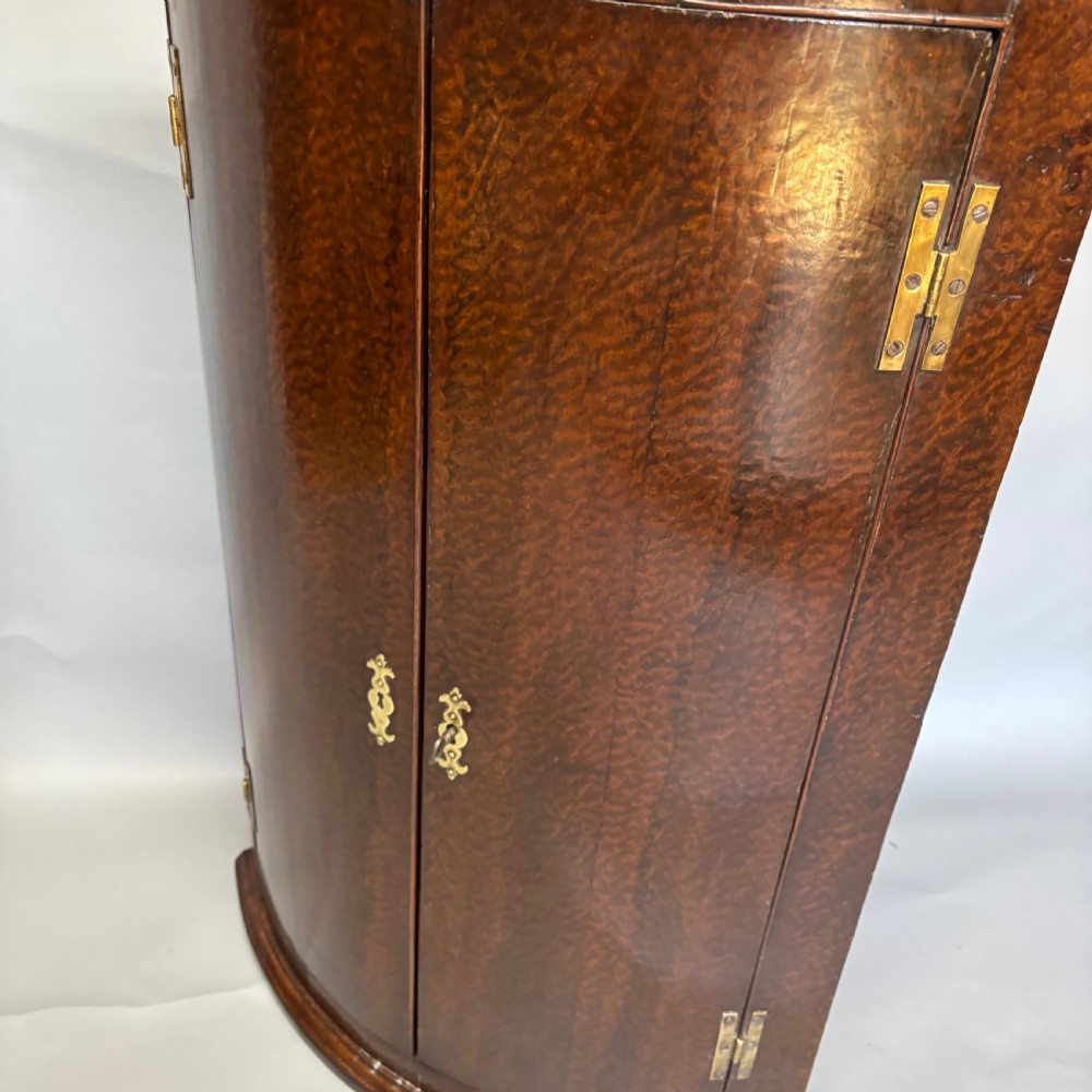 A Georgian bowfront mahogany corner cupboard featuring distinctive plum pudding grain, brass escutcheons, and visible brass H-hinges. View VI
