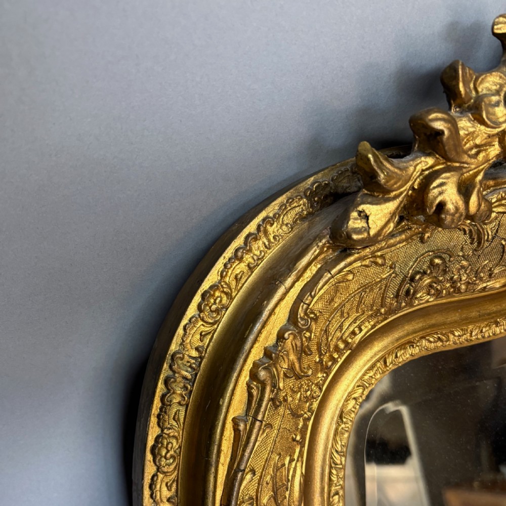A 19th-century French giltwood wall mirror featuring an intricate floral cresting, molded frame, and carved scroll corner accents. View V