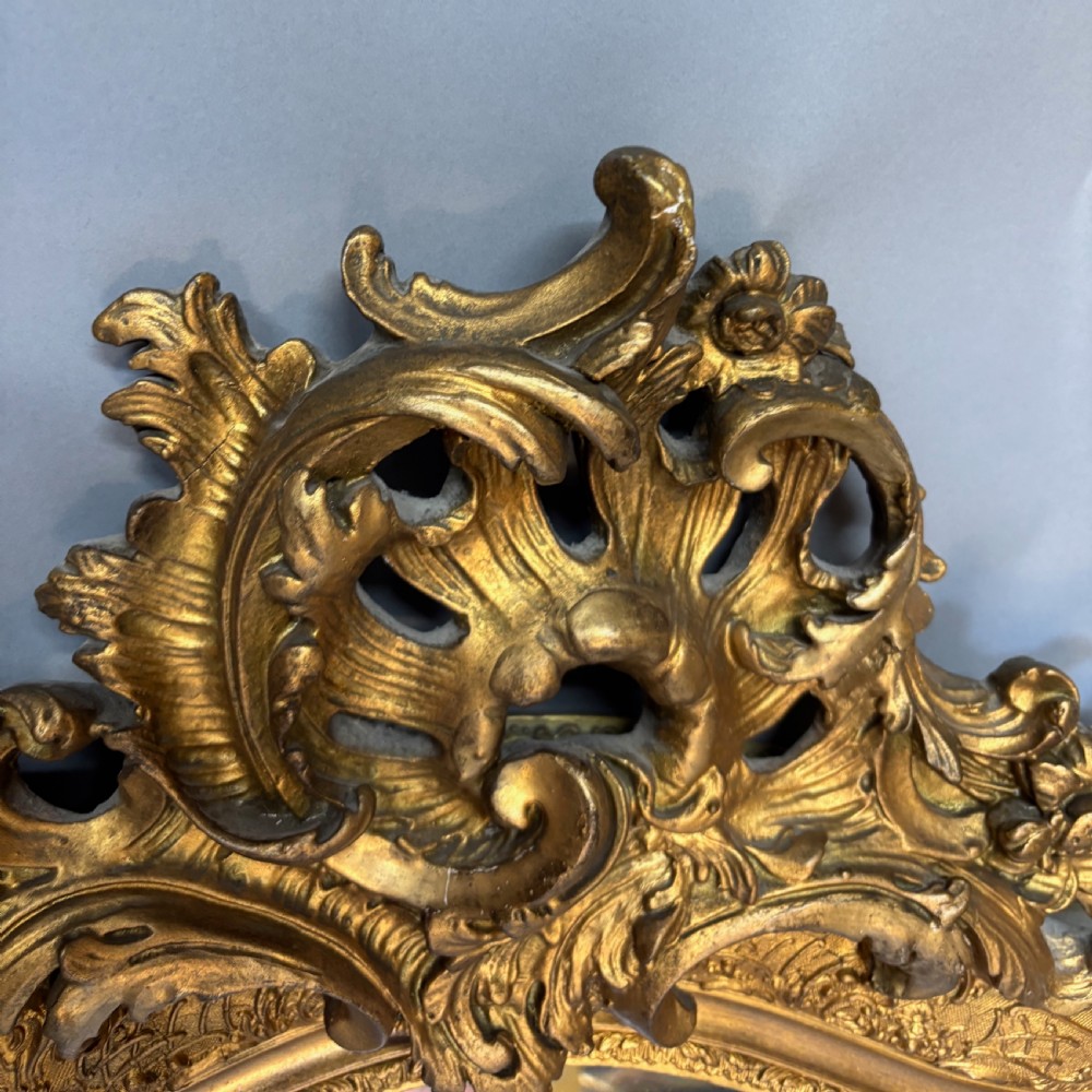 A 19th-century French giltwood wall mirror featuring an intricate floral cresting, molded frame, and carved scroll corner accents. View VI