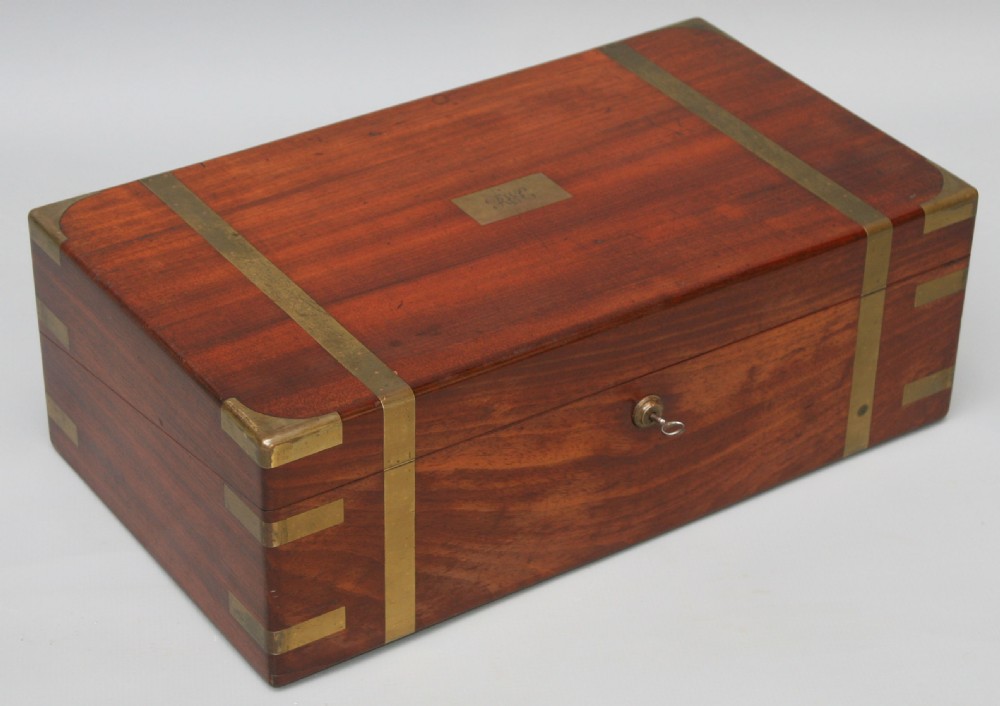 A rectangular early Victorian mahogany writing box featuring flush brass binding, brass corner plates, a central brass plaque, and a brass keyhole with a key inserted. View II