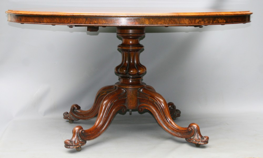 An oval Victorian burr walnut loo table featuring a figured top resting on a central carved pedestal with four splayed legs. View III