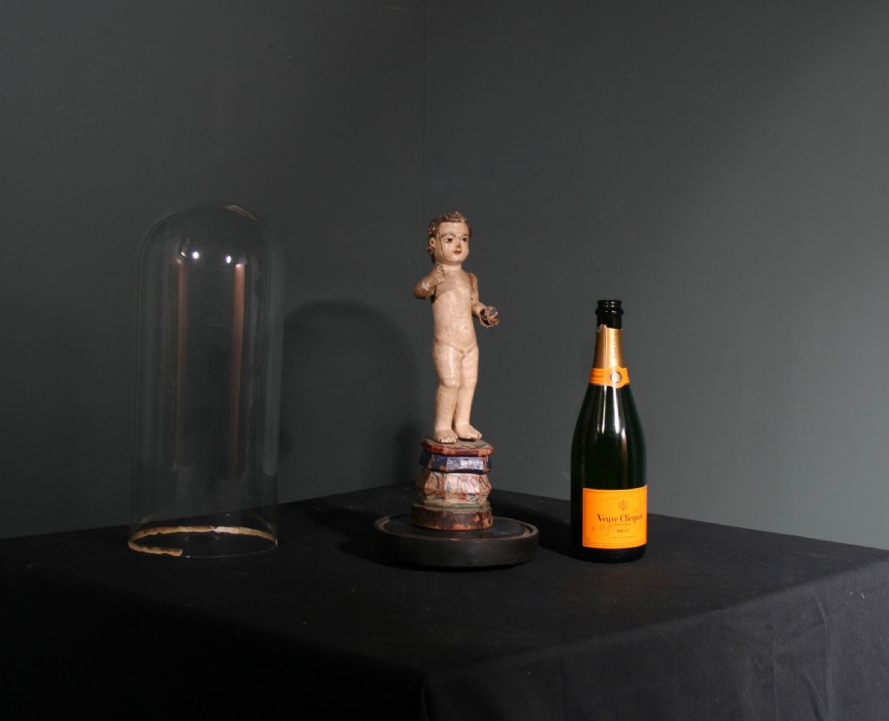 A carved wooden figure of the Christ Child holding an orb, mounted on a painted faceted base and displayed beneath a clear glass cloche. View II